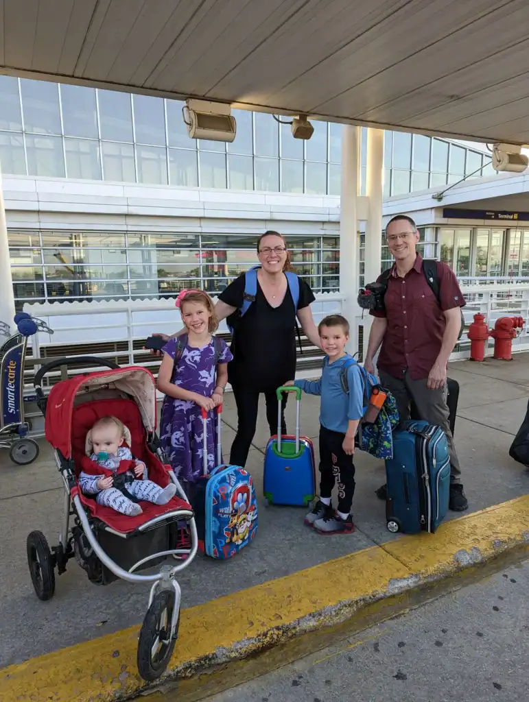 A smiling family of five stands outside an airport terminal with rolling suitcases and a stroller, ready to kick off their next adventure. This candid travel moment reflects the heart of their family travel blog.